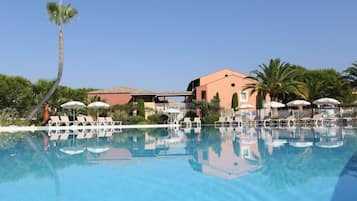 Une piscine d’un bleu éclatant, bordée de chaises longues et d’ombrelles, entourée de palmiers et d’un bâtiment dont le toit est recouvert de tuiles.