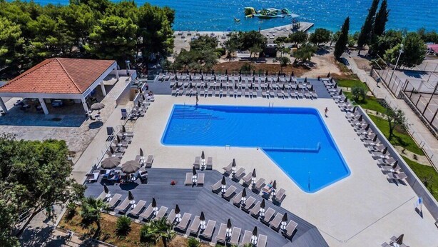 A large swimming pool with sun loungers and a covered area, surrounded by trees and a view of the sea. - Hotel Medena Block A (Seget)