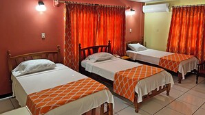 A hotel room with three single beds, each with a white and orange patterned bedspread. The room has red walls, a tiled floor, and an air conditioning unit on the wall.