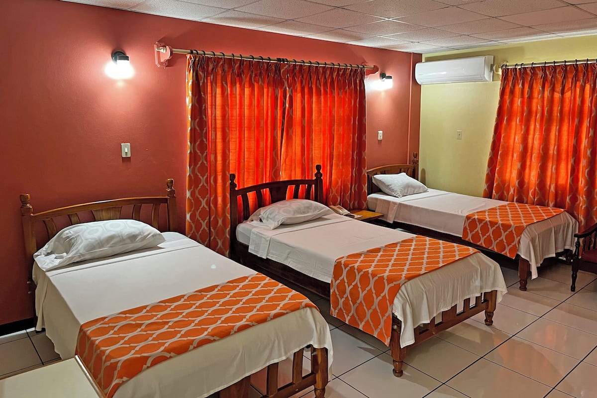 Habitación de hotel con tres camas individuales, cada una con colcha blanca y anaranjada. La habitación tiene paredes rojas, piso de baldosas y un equipo de aire acondicionado en la pared.