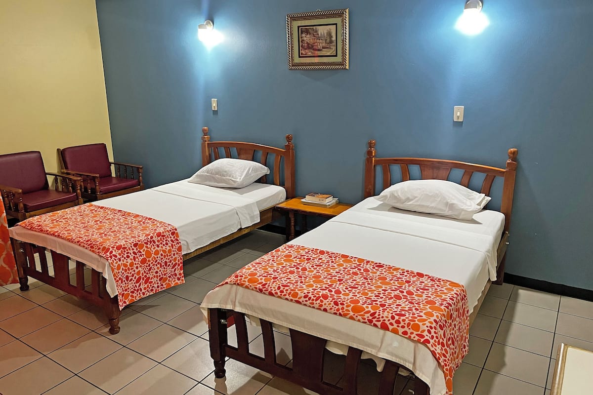 Habitación de hotel con dos camas individuales, una mesita, dos sillas y un cuadro en la pared.