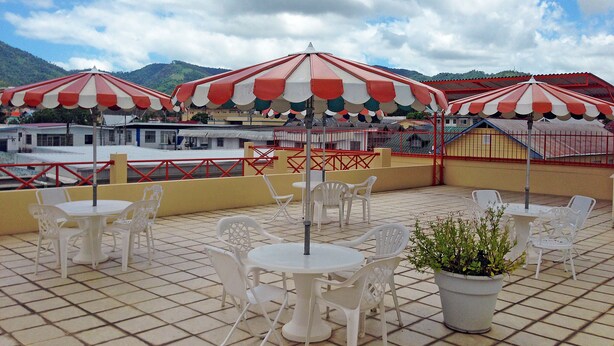 A terrace with white plastic chairs and tables, red and white striped umbrellas, and a view of a mountainous landscape.