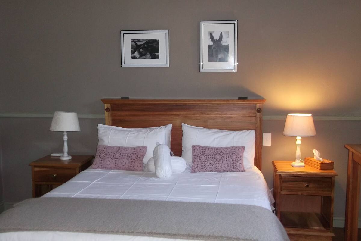 A neatly made bed with white and purple pillows, a wooden headboard, and a bedside table with a lamp.