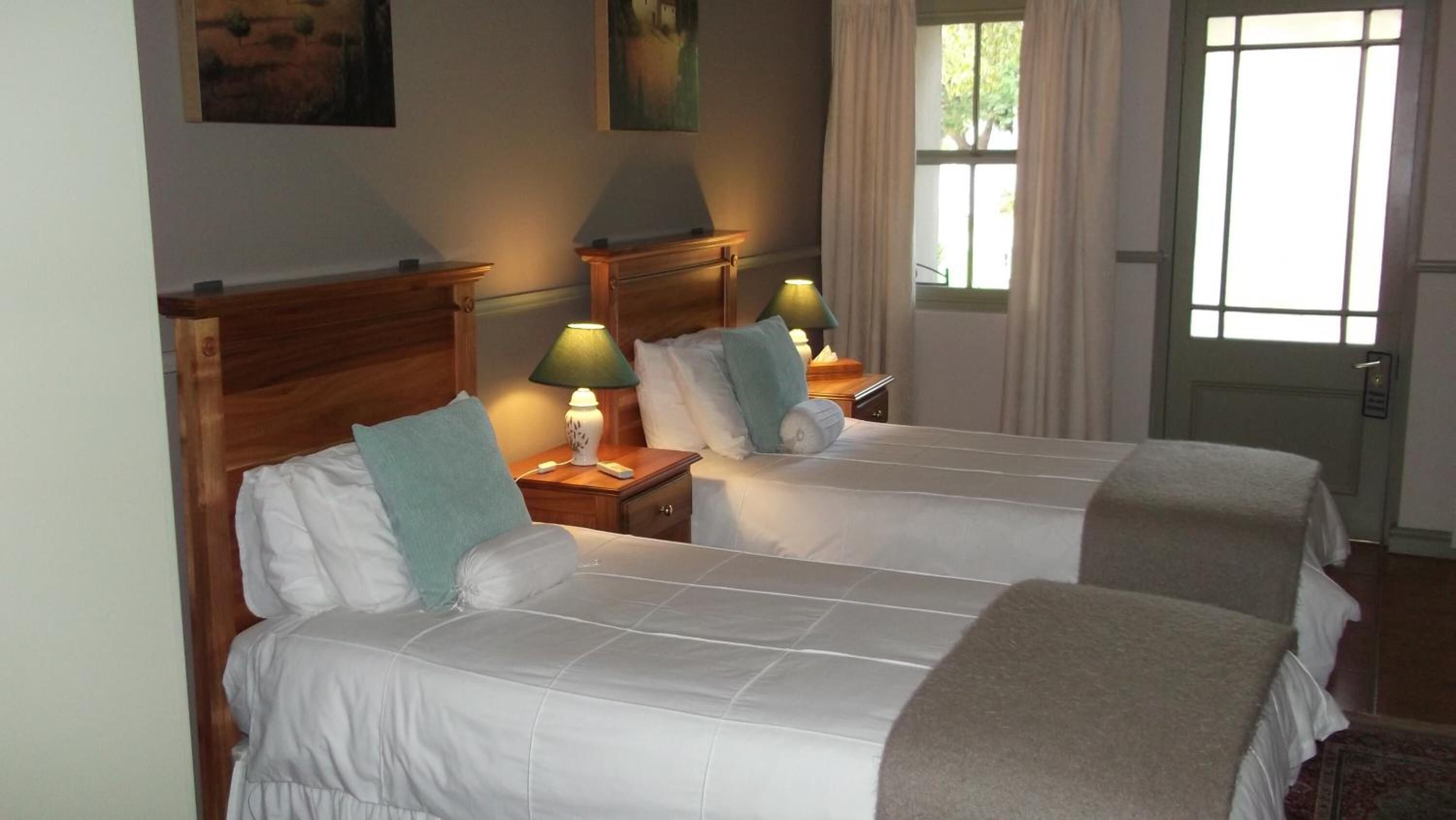Camera d'albergo con due letti, ognuno con testiera in legno, biancheria da letto bianca e cuscino verde. Sono presenti comodini con lampade e una finestra con tende.
