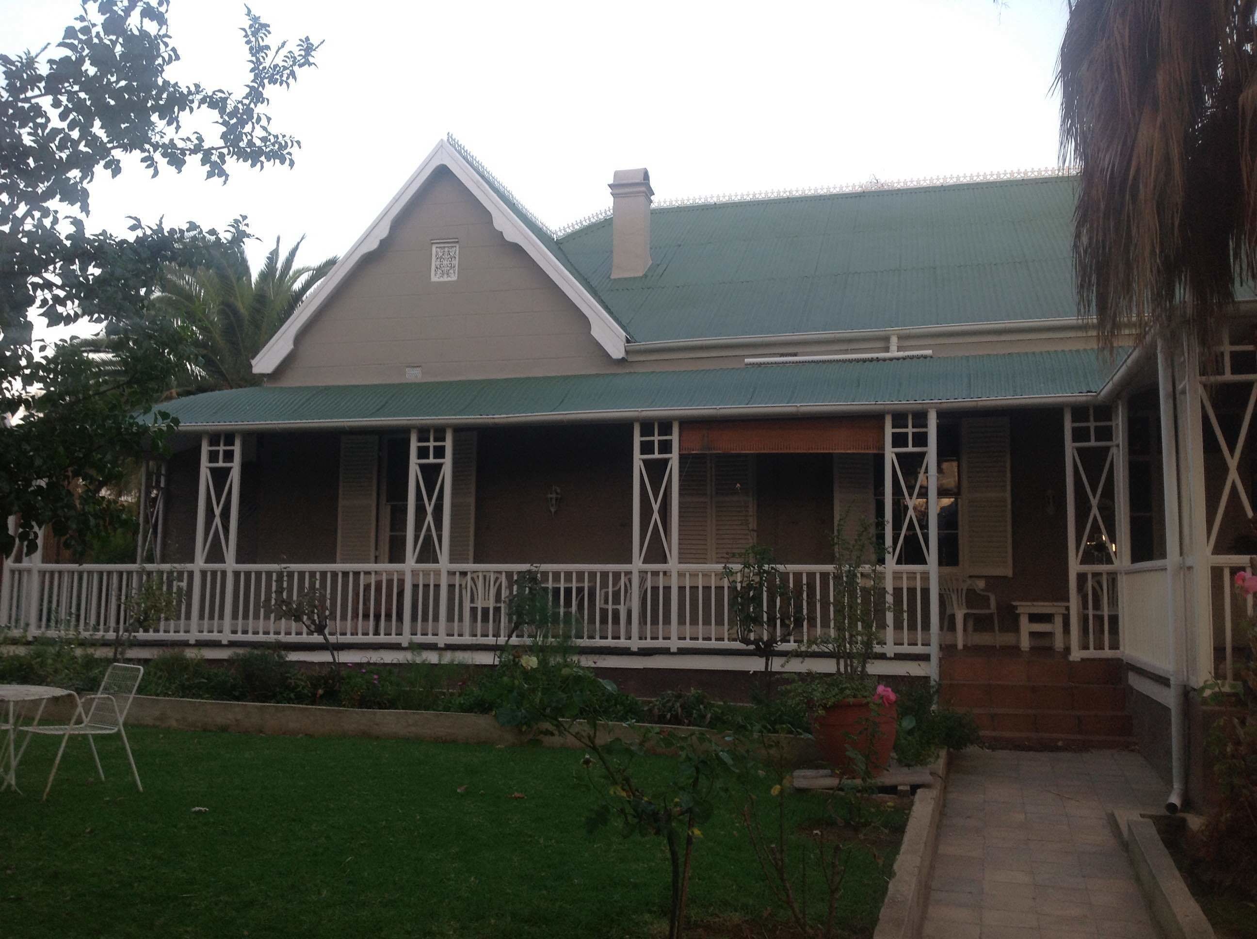 Uma casa de dois andares com telhado verde, varanda com grades brancas e um pequeno jardim com gramado e plantas em vasos.