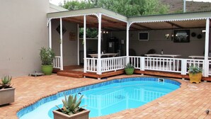 Une piscine avec une terrasse en bois, un patio couvert doté de rampes blanches et des plantes en pot.