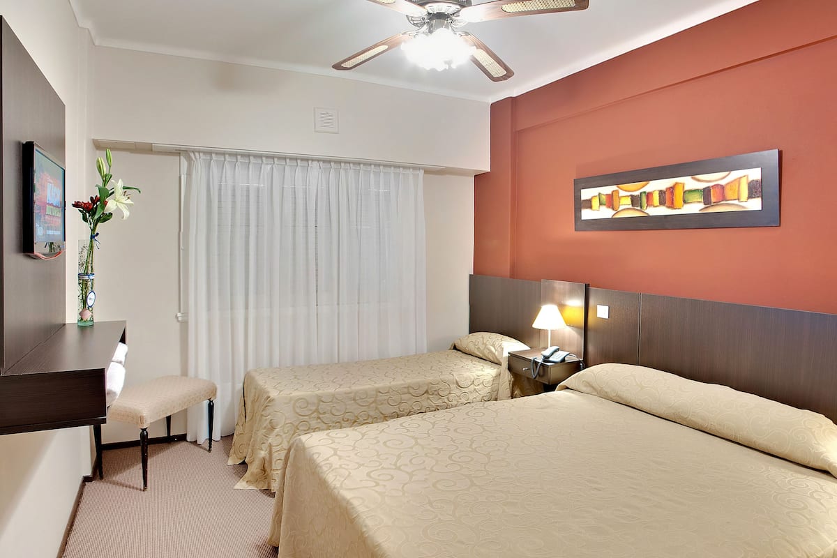 Hotelkamer met twee bedden, een bureau, een stoel, een plafondventilator, een schilderij en een nachtkastje met een lamp.