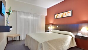 Een hotelkamer met een bed, nachtlampjes, een tafeltje met een telefoon en lamp, een bankje, een vaas met bloemen en een schilderij aan de muur.