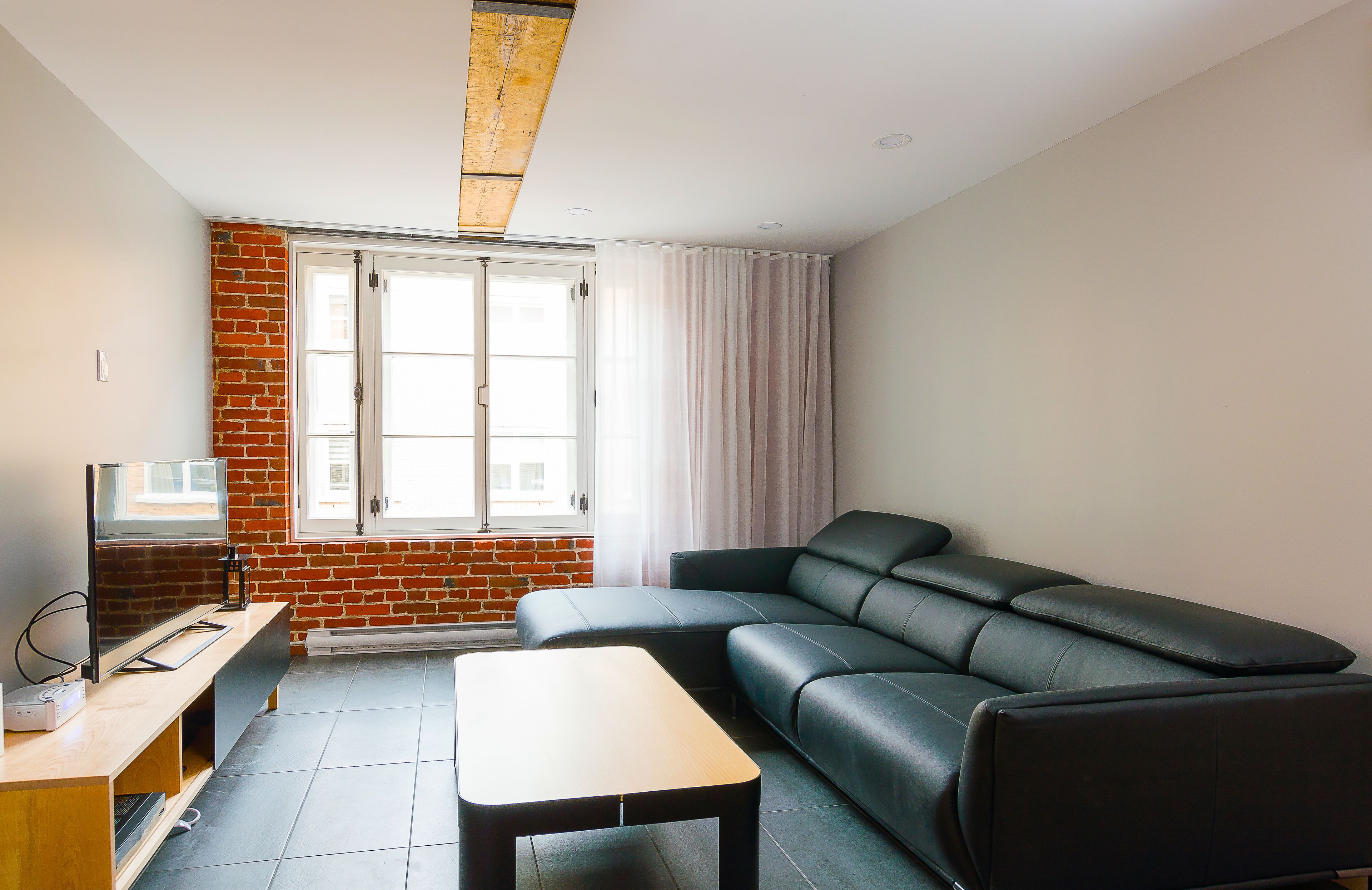 A modern living room with a black leather sofa, a wooden coffee table, and a TV on a wooden stand. A brick wall and a window with curtains are visible.