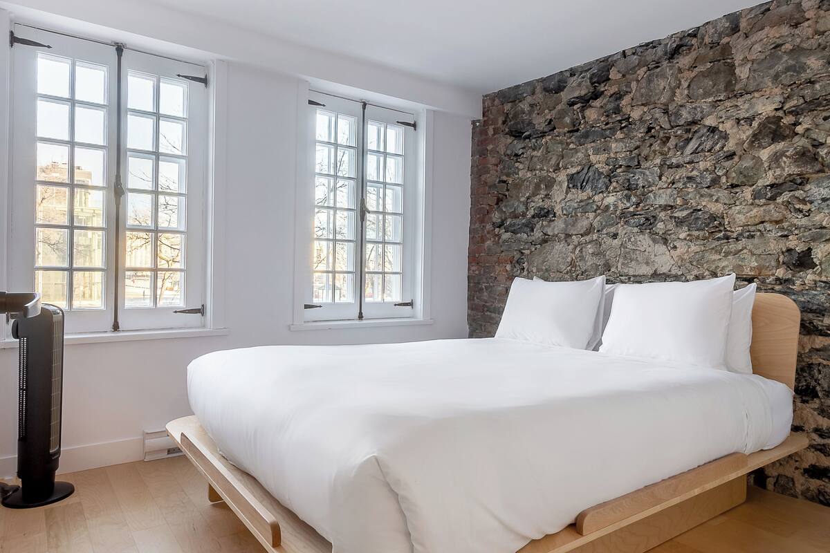 A modern bedroom with a large bed, two windows, a stone wall, and a standing heater.