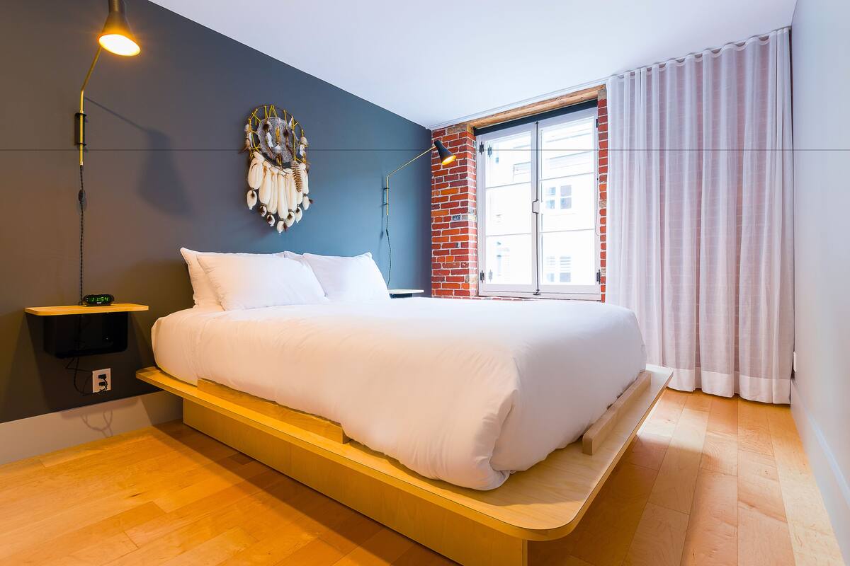A modern bedroom with a bed, a nightstand, a wall-mounted lamp, a dreamcatcher, a brick wall, and a window with curtains.