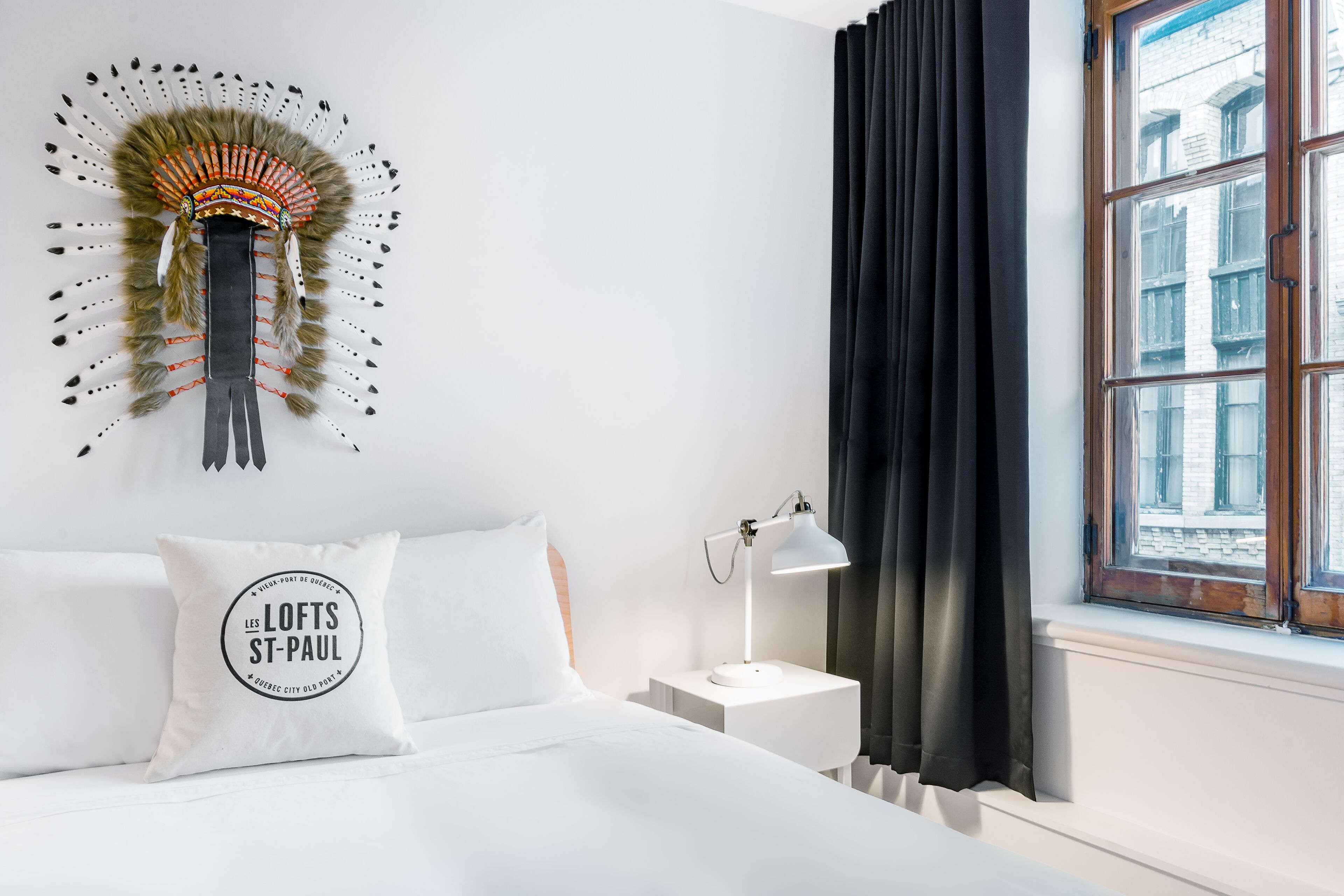 A hotel room with a bed, a nightstand, a lamp, a window with curtains, and a decorative wall piece.
