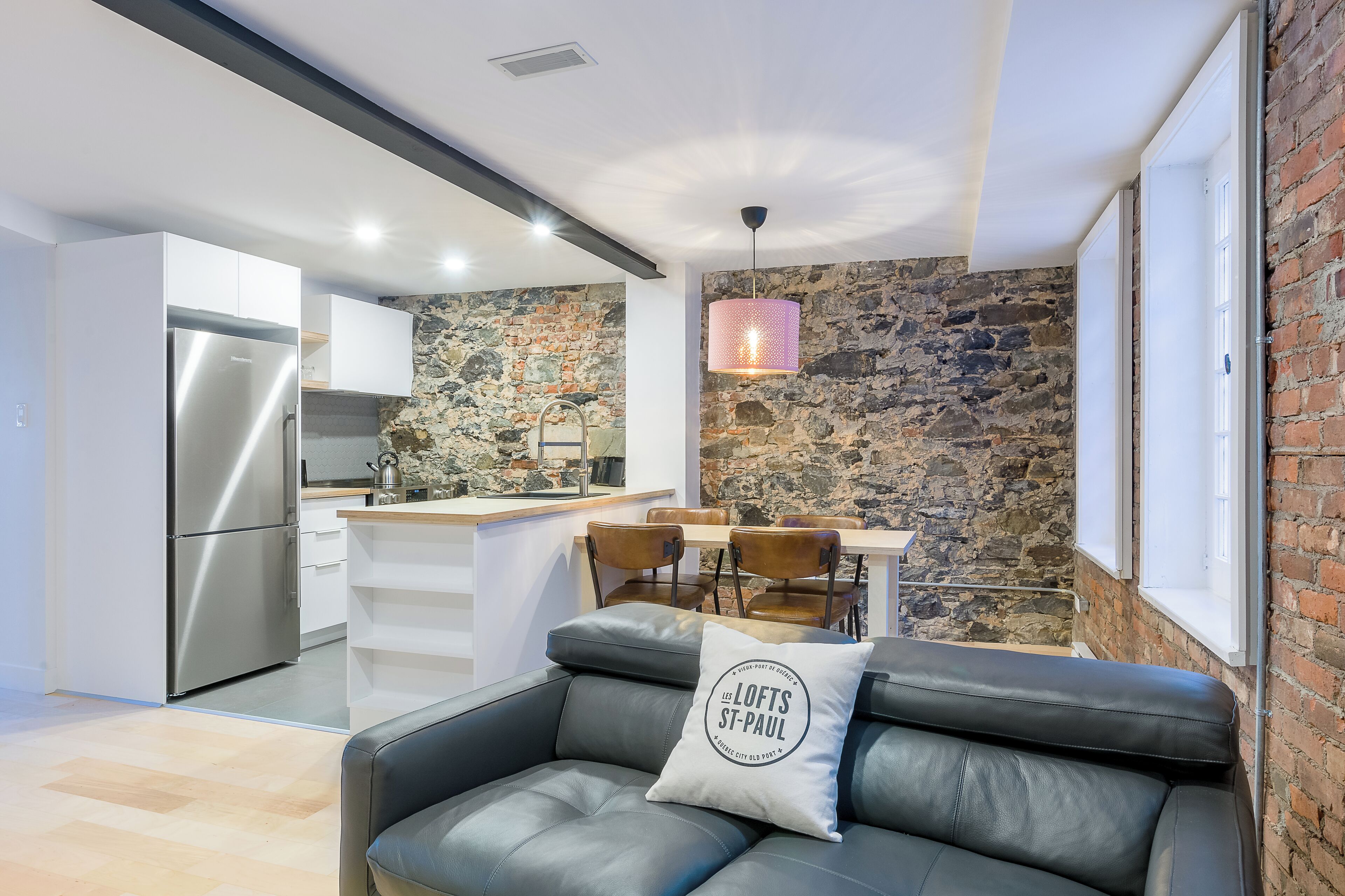 Une cuisine moderne avec un mur en pierre, un coin repas avec une table et des chaises, et un canapé en cuir noir agrémenté d’un coussin.