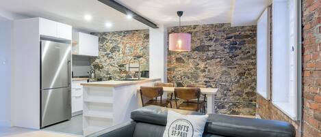 Une cuisine moderne avec un mur en pierre, un coin repas avec une table et des chaises, et un canapé en cuir noir agrémenté d’un coussin.