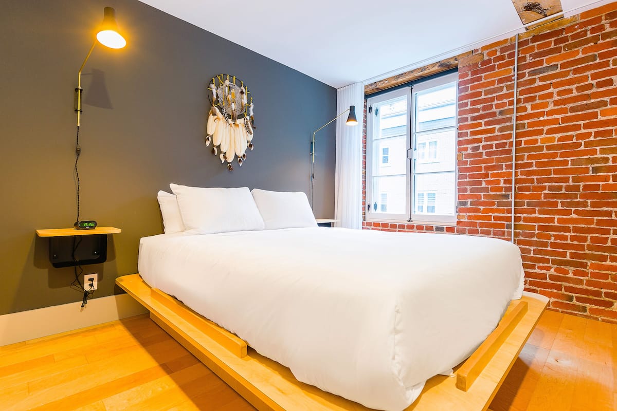 A modern bedroom with a wooden platform bed, a wall-mounted nightstand, a decorative wall piece, and a brick wall.