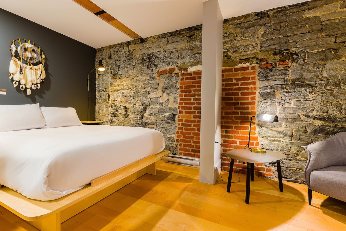 A bedroom with a bed, a nightstand, a lamp, a chair, and a stone wall.