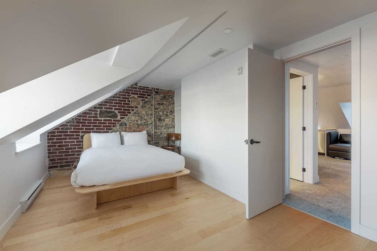 A modern bedroom with a lofted sleeping area, a brick accent wall, and a view of the living area through an open door.