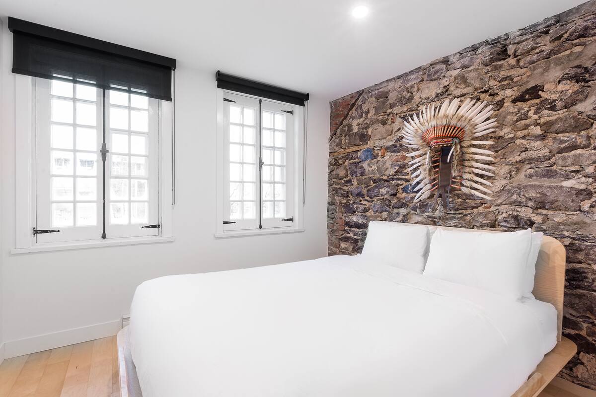 A bedroom with a large bed, two windows with black blinds, a stone wall, and a decorative wall piece.