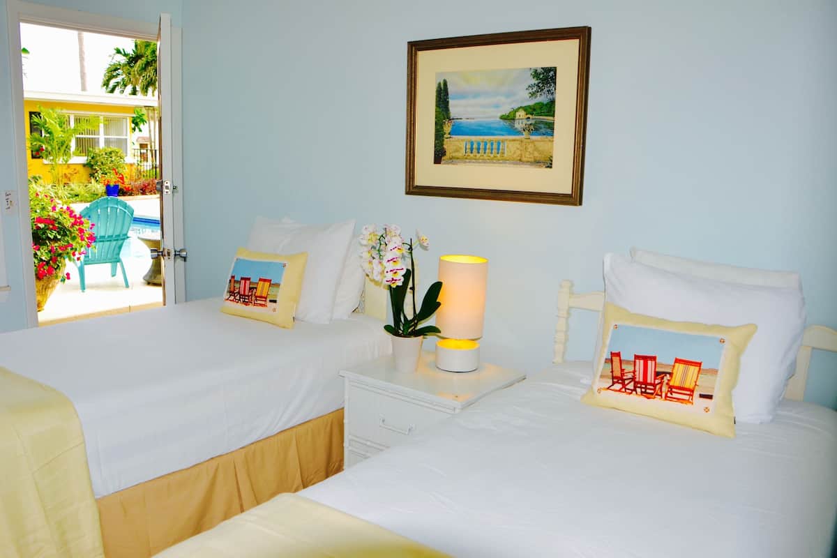 A hotel room with two beds, a nightstand with a lamp, a framed picture on the wall, and a view of the outside balcony.