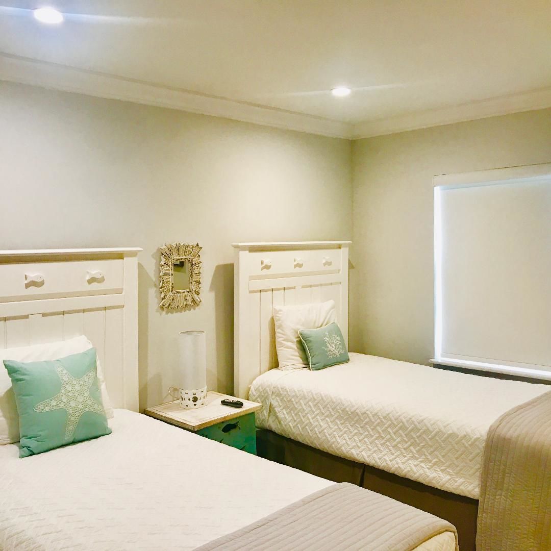 A room with two beds, each with a white headboard and a single pillow. There is a small bedside table with a lamp and a mirror on the wall.