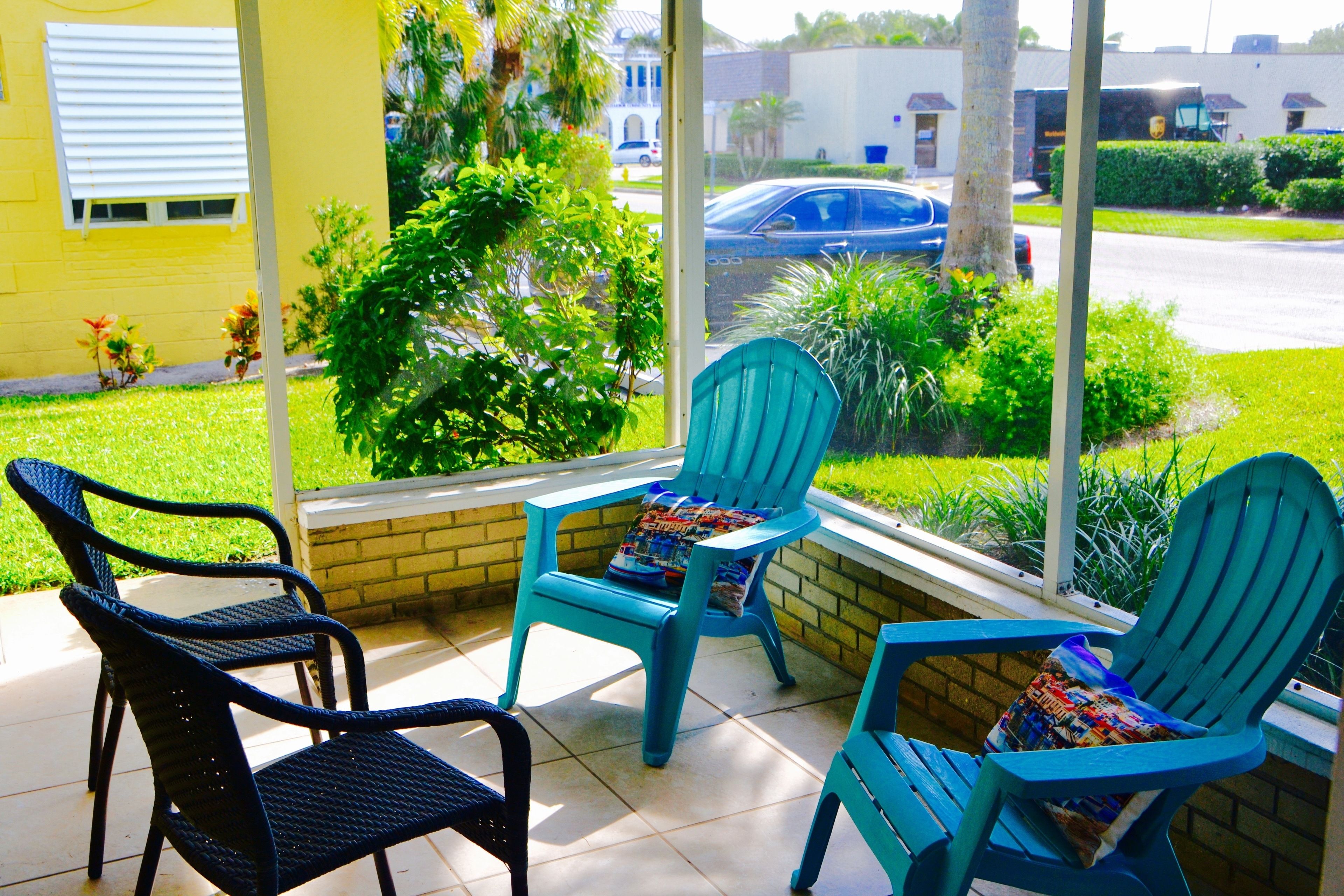 A patio with two blue Adirondack chairs and a wicker chair, overlooking a street with cars and greenery.