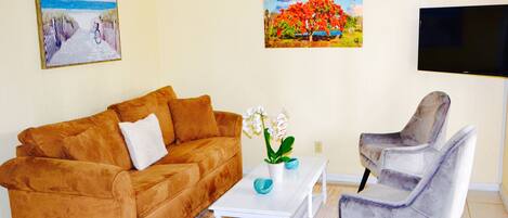 A living room with a brown sofa, a white coffee table with a vase of flowers, a grey armchair, and two wall-mounted paintings.