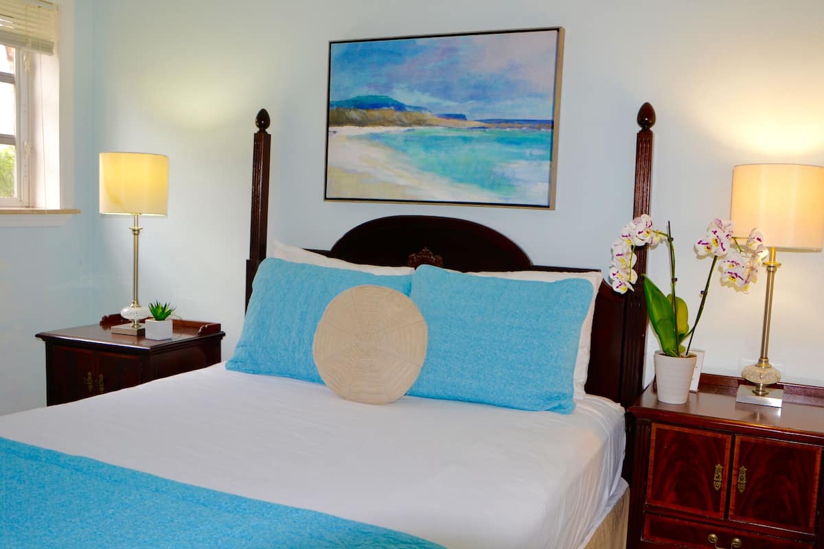 A neatly made bed with blue and beige pillows, a painting of a beach scene, and two bedside tables with lamps and a potted orchid.