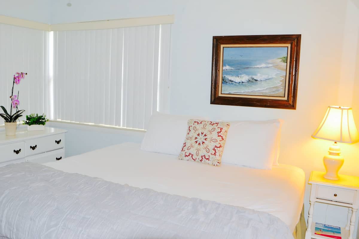 A hotel room with a bed, a nightstand with a lamp, a framed picture on the wall, and a window with blinds.
