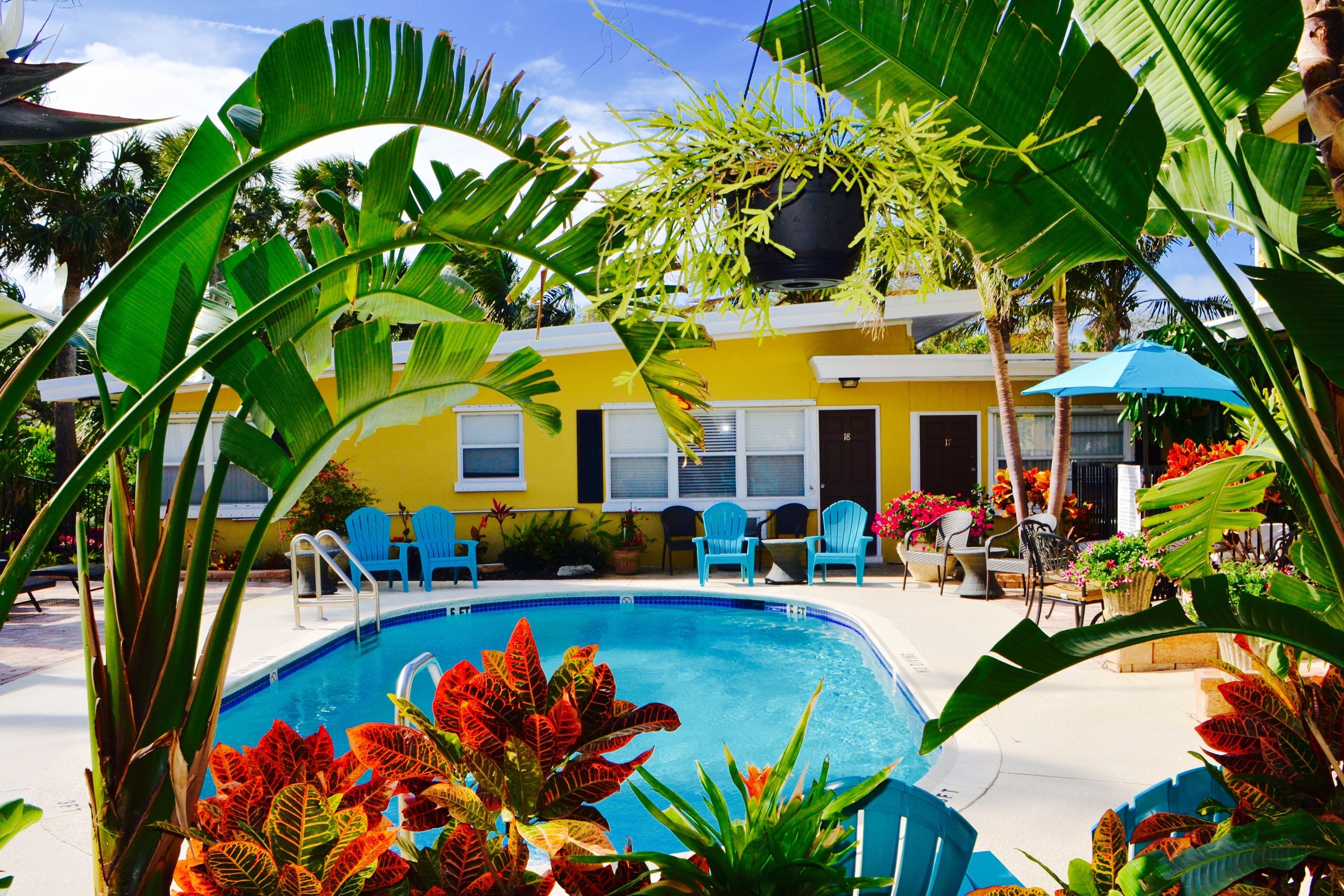 A pool area with a yellow building, blue chairs, and tropical plants.