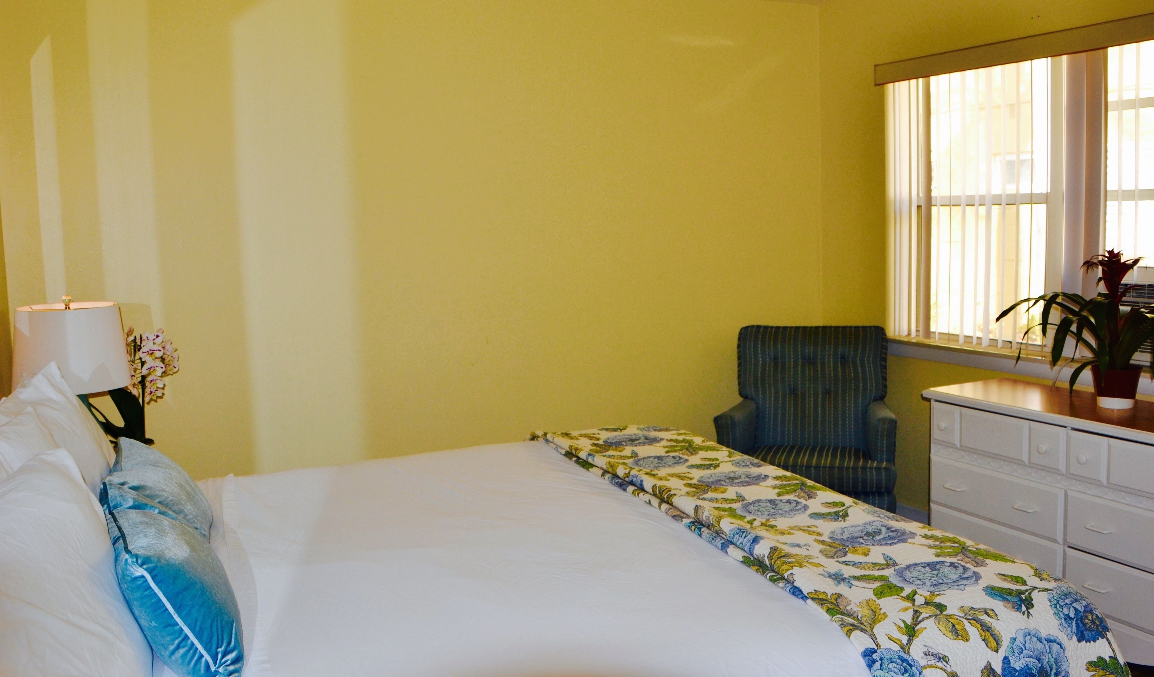 Une chambre d’hôtel avec un lit, une chaise, une commode et une fenêtre avec des stores.