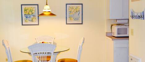 Un comedor con mesa de vidrio y sillas de mimbre blanco, lámpara colgante y cuadros en la pared.