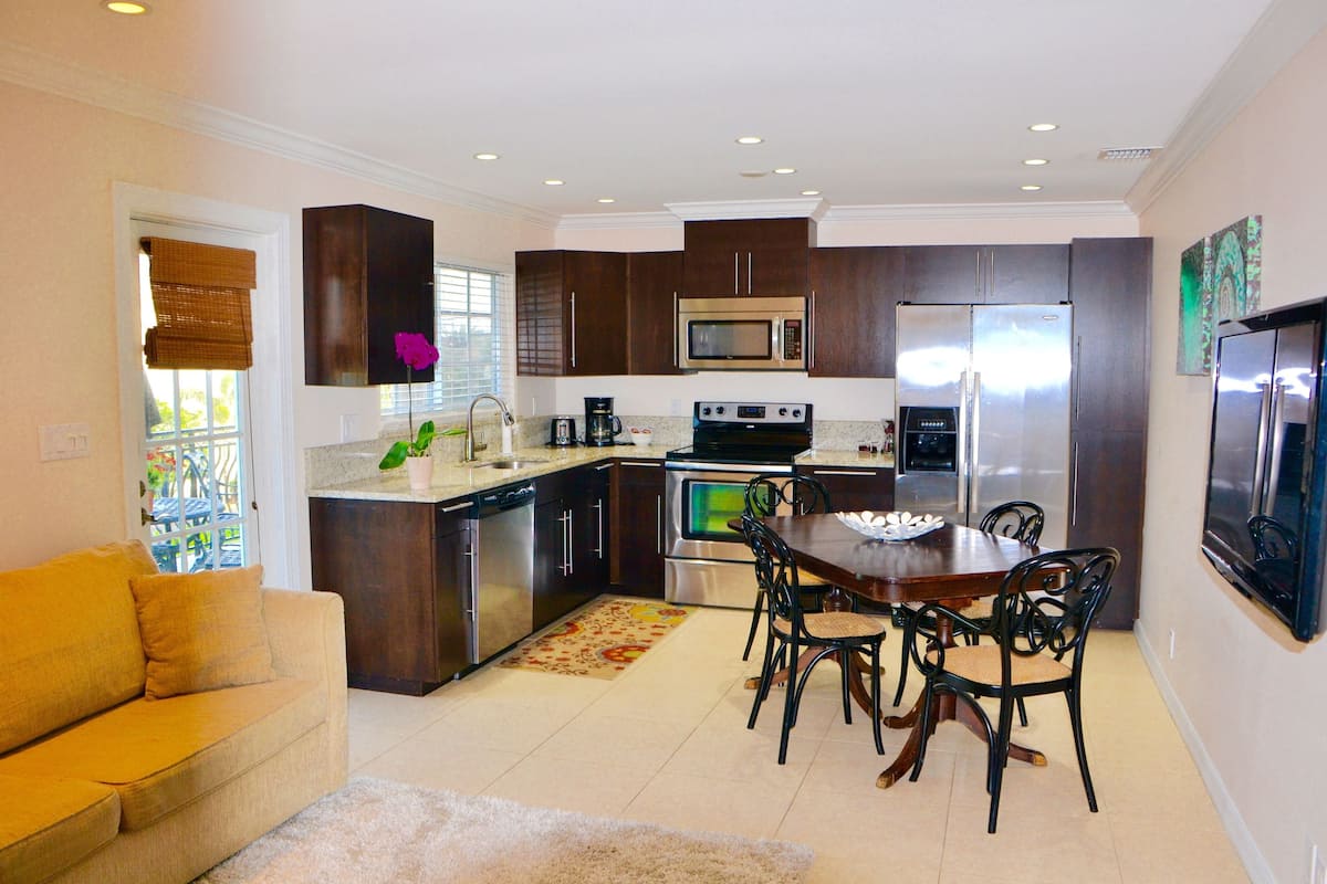 A modern kitchen with dark cabinets, stainless steel appliances, a dining area with a wooden table and chairs, and a yellow sofa.