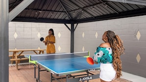 Una mesa de ping pong en el interior con dos personas jugando, una pared de ladrillo con diseños de diamante y un techo metálico.