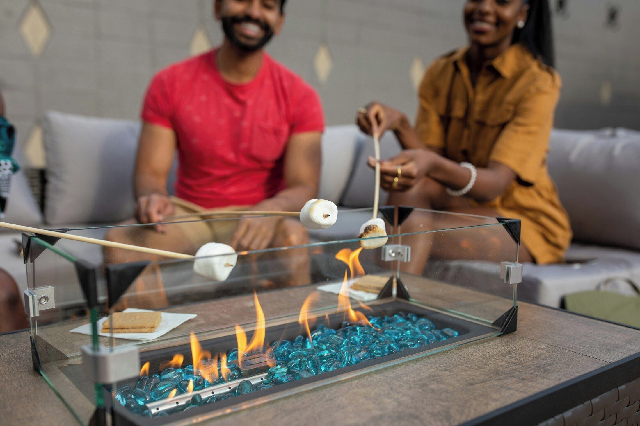 Un foyer avec un dessus en verre, agrémenté de petits galets de verre bleus, un porte-bâtons en bois pour les chamallows, et une personne en train de faire griller des chamallows.