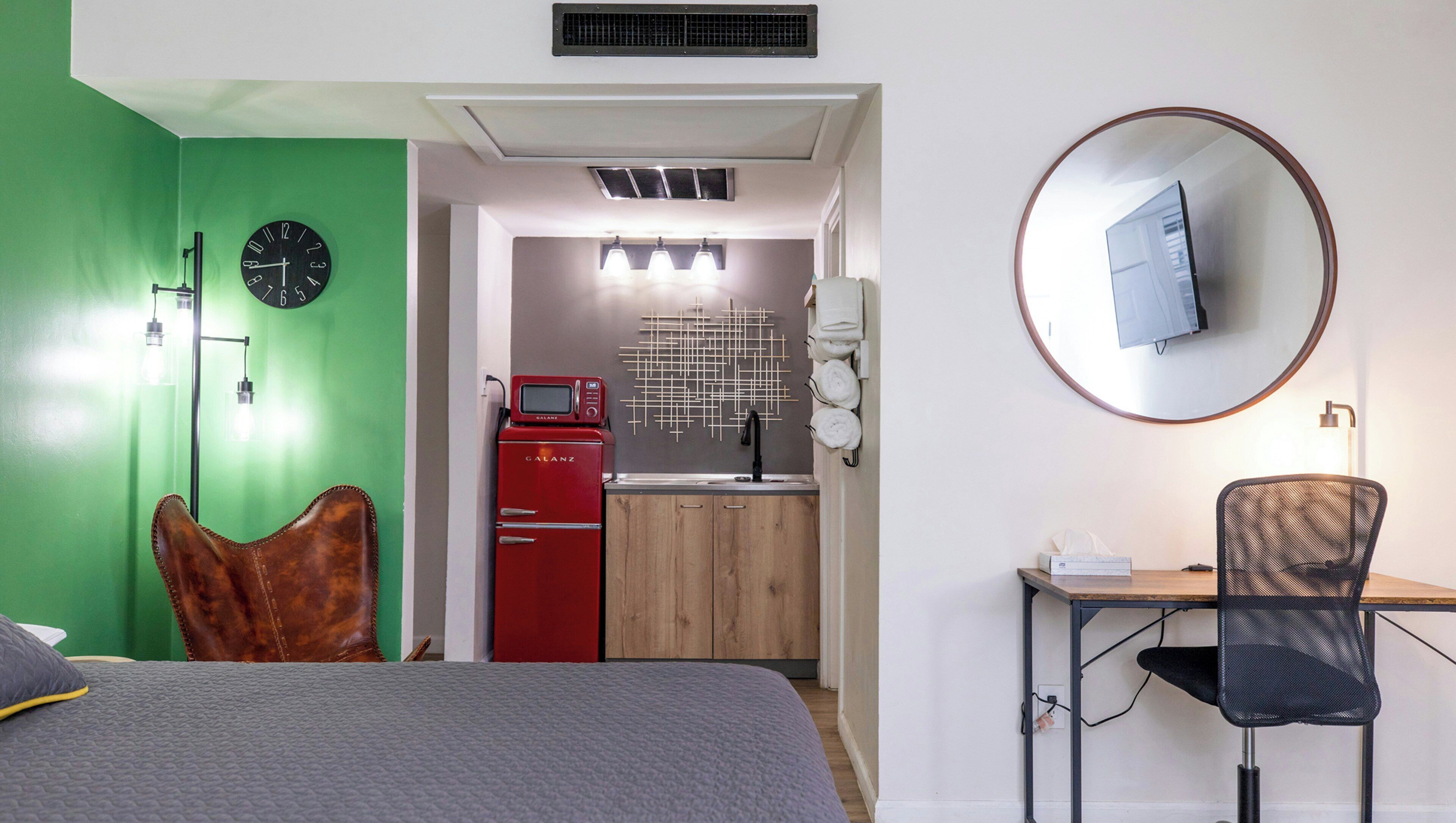 Une chambre d’hôtel comprenant un lit, un bureau avec une chaise, un coin cuisine, un miroir et un mur vert.