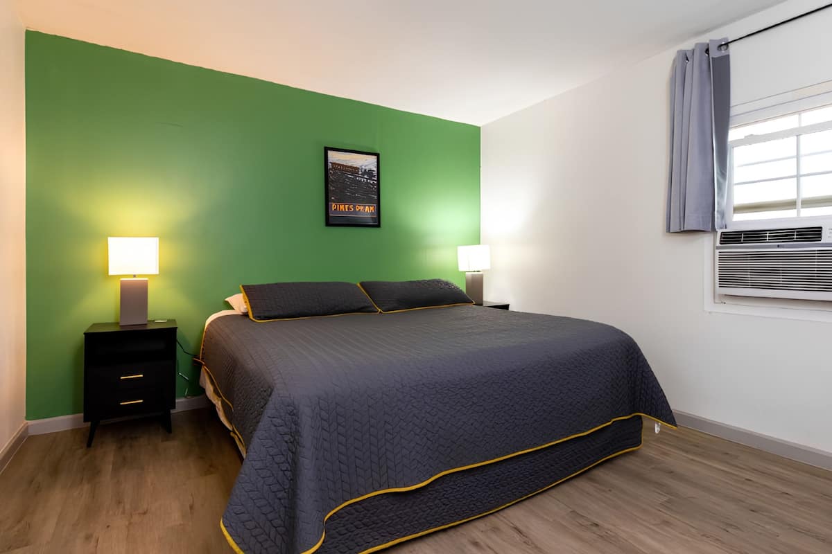 Une chambre à coucher comprenant un lit, deux tables de chevet, un climatiseur fixé au mur et une fenêtre avec des rideaux.