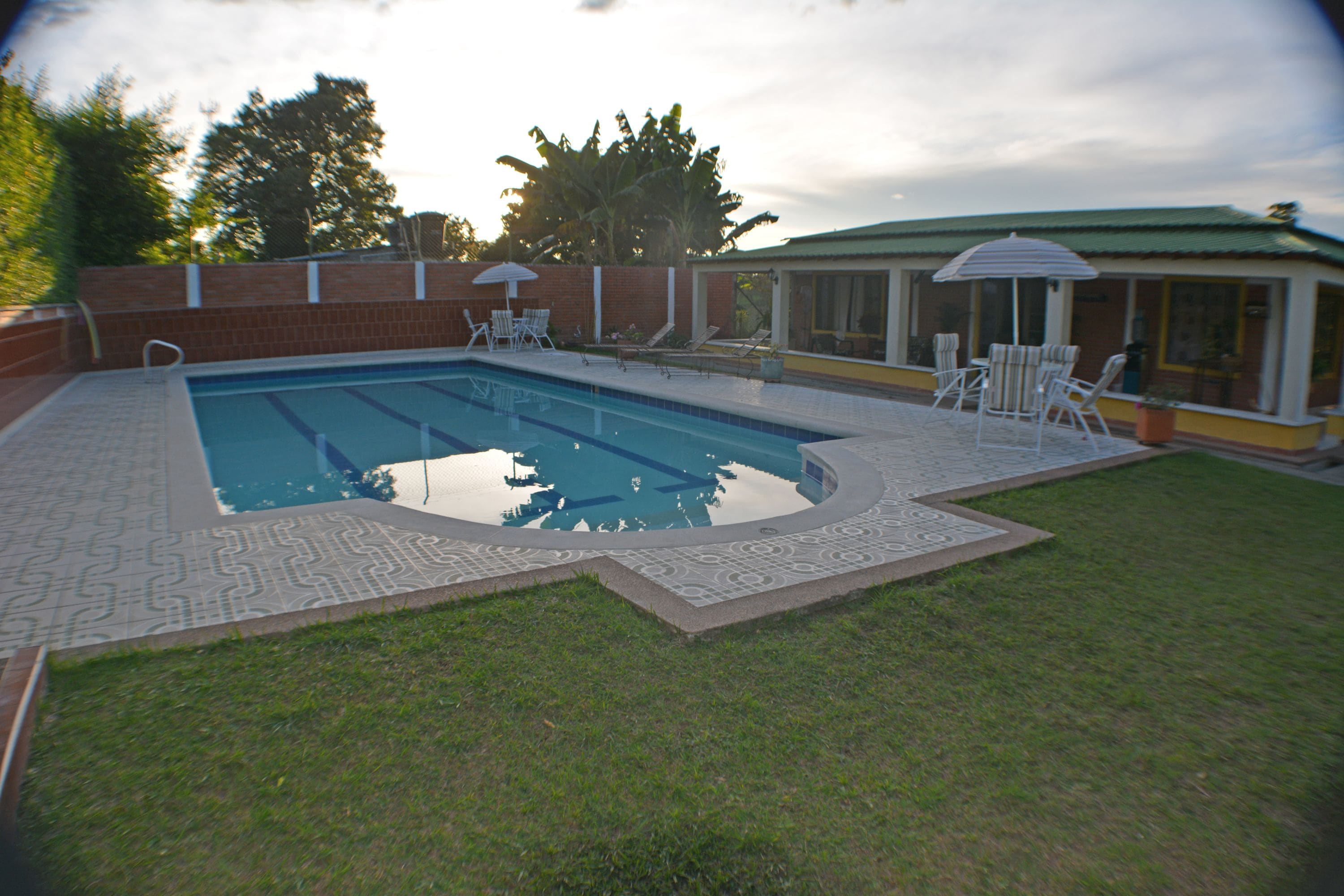 Une piscine rectangulaire bordée d’une terrasse carrelée, entourée d’un mur de briques et agrémentée d’un patio couvert avec des chaises et un parasol.