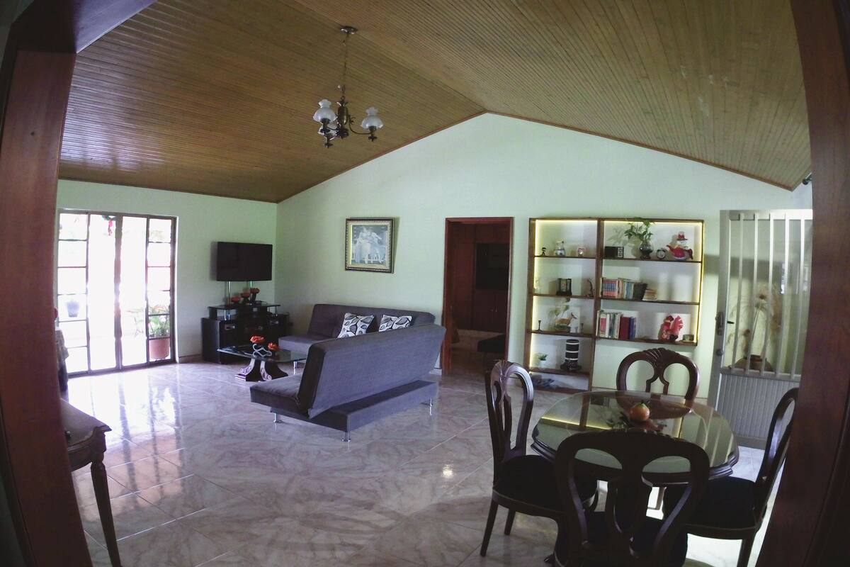 A spacious living area with a high ceiling, a sofa, a dining table with chairs, and a bookshelf.