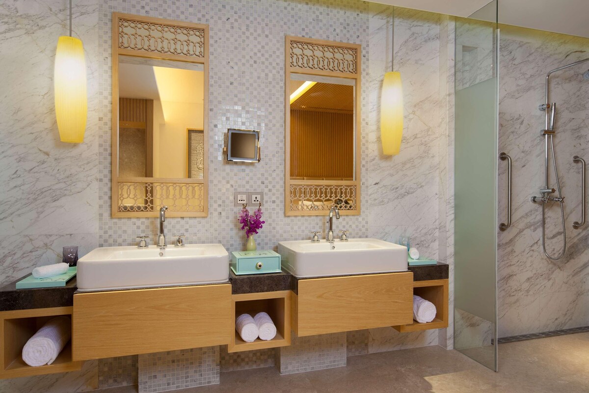 Een moderne badkamer met twee wastafels, een houten wastafelmeubel, een douchecabine met glazen wanden en decoratieve spiegels.