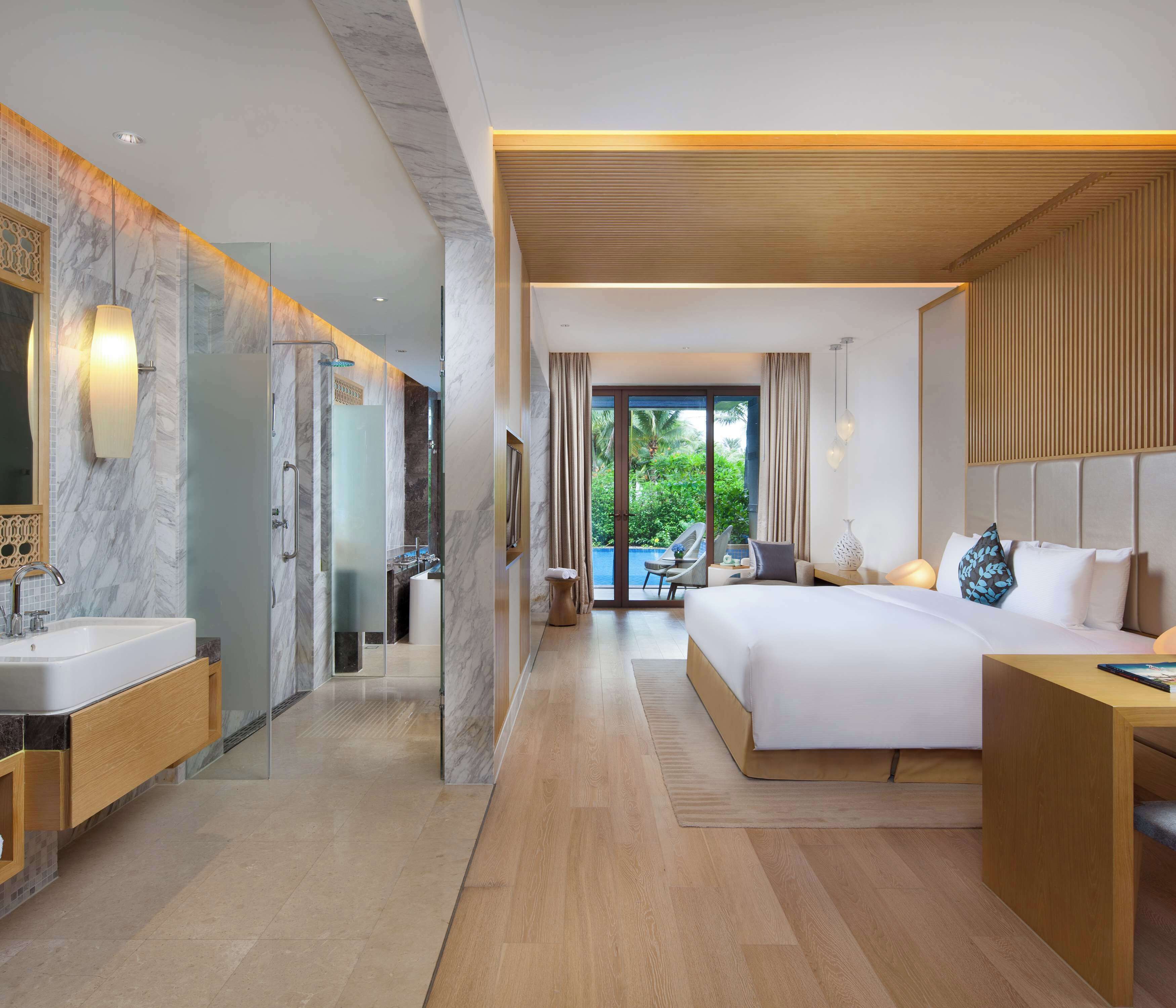 A modern hotel room with a large bed, a bathroom with a glass shower, and a view of greenery outside.