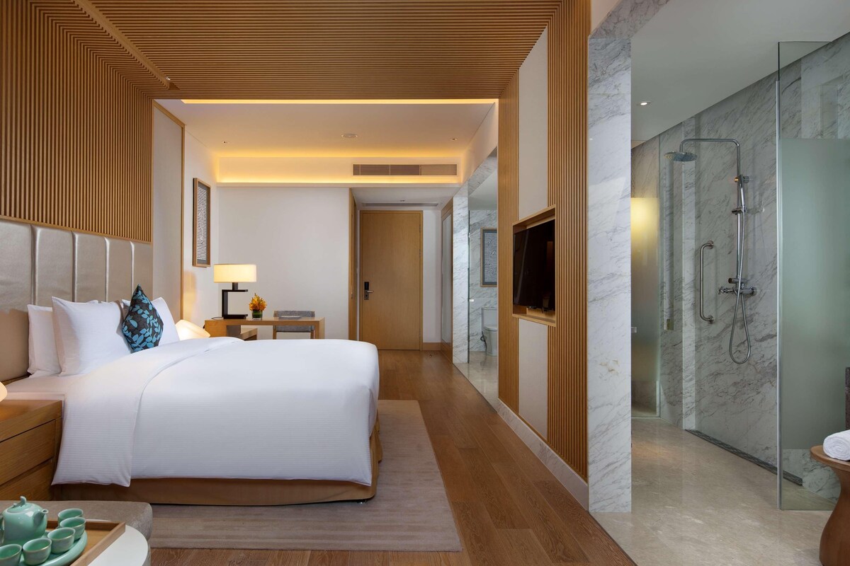 Een moderne hotelkamer met een groot bed, een nachtkastje met een lamp, een televisie en een douchecabine met glazen wanden.