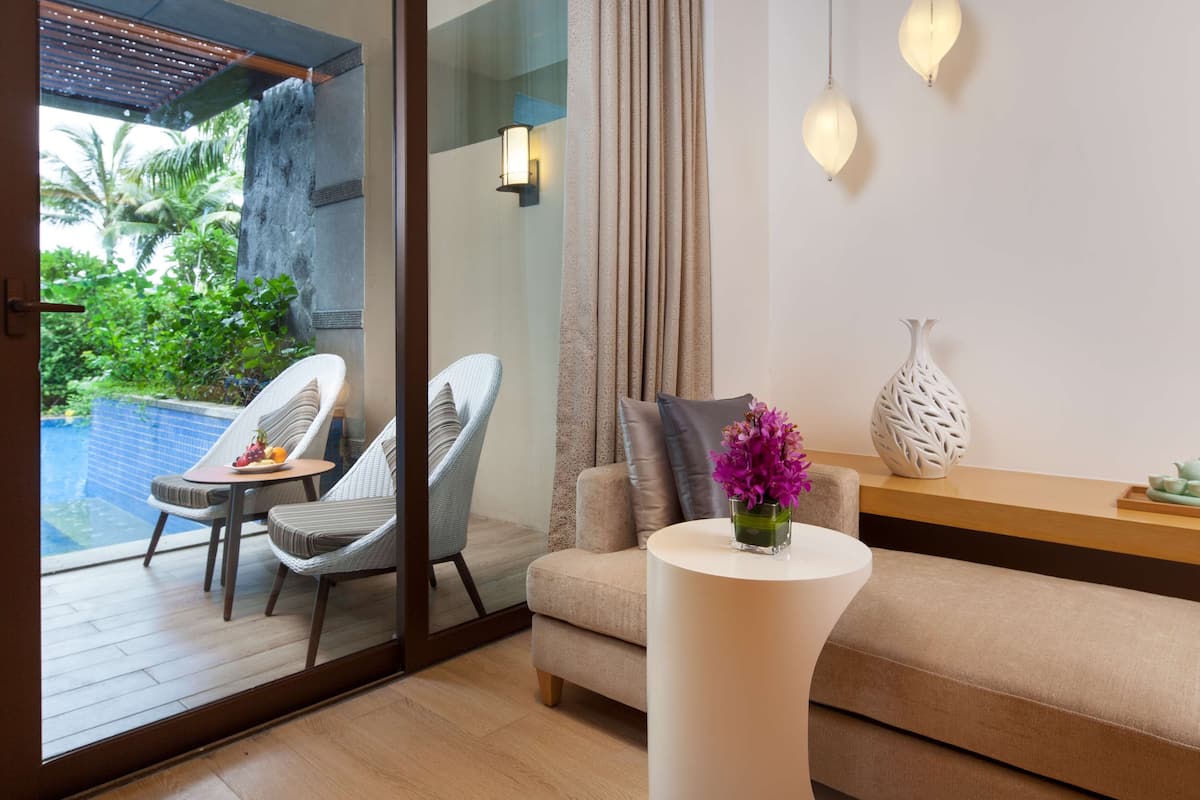 A modern living room with a sofa, a coffee table, and two chairs. There is a glass door leading to a balcony with a pool.