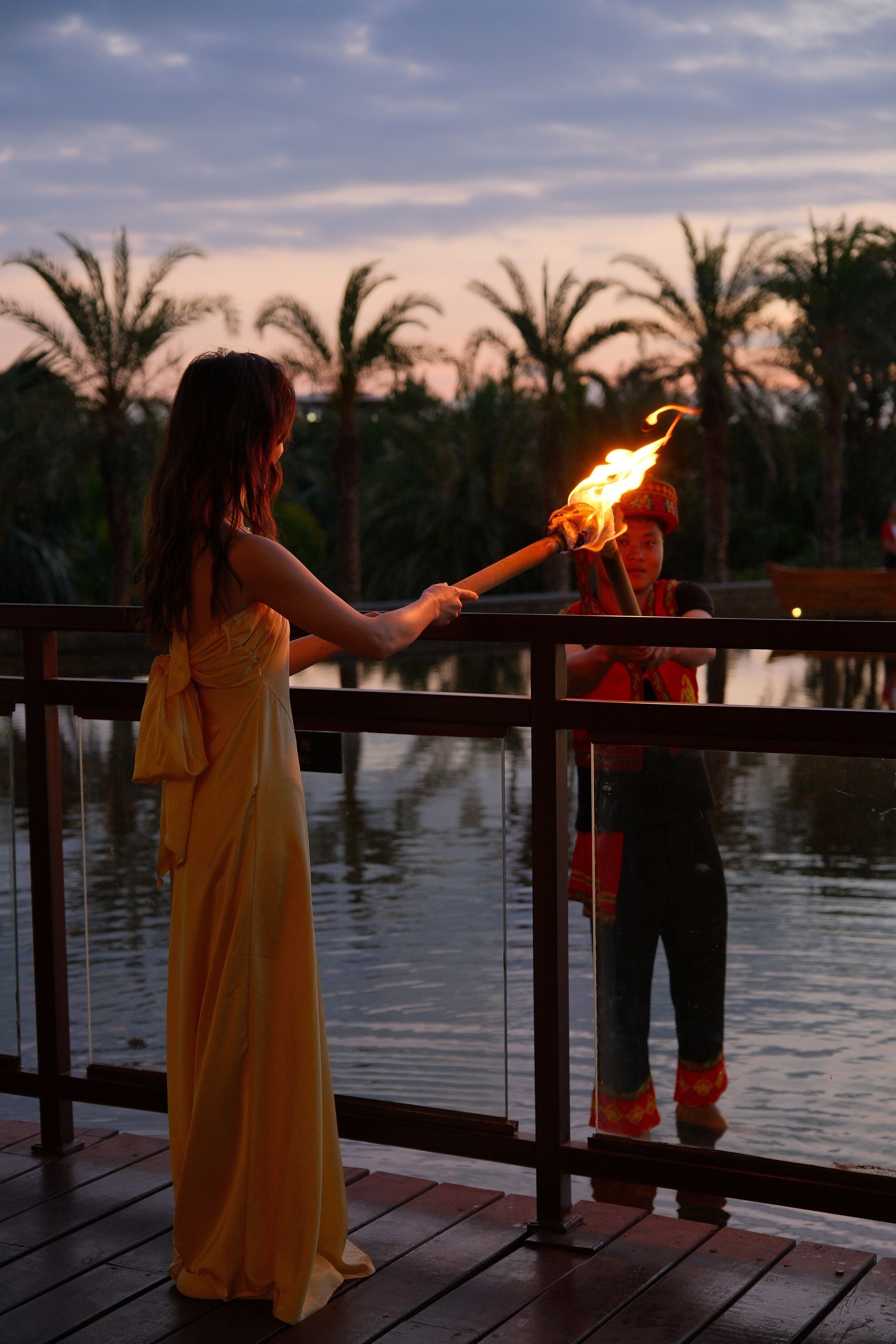Una persona con un vestido amarillo sosteniendo una antorcha, de pie sobre una cubierta de madera con barandilla, con vistas a un cuerpo de agua y palmeras.