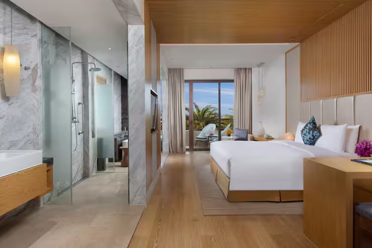 Een moderne hotelkamer met een groot bed, een houten bureau, een badkamer met douche en uitzicht op palmbomen.