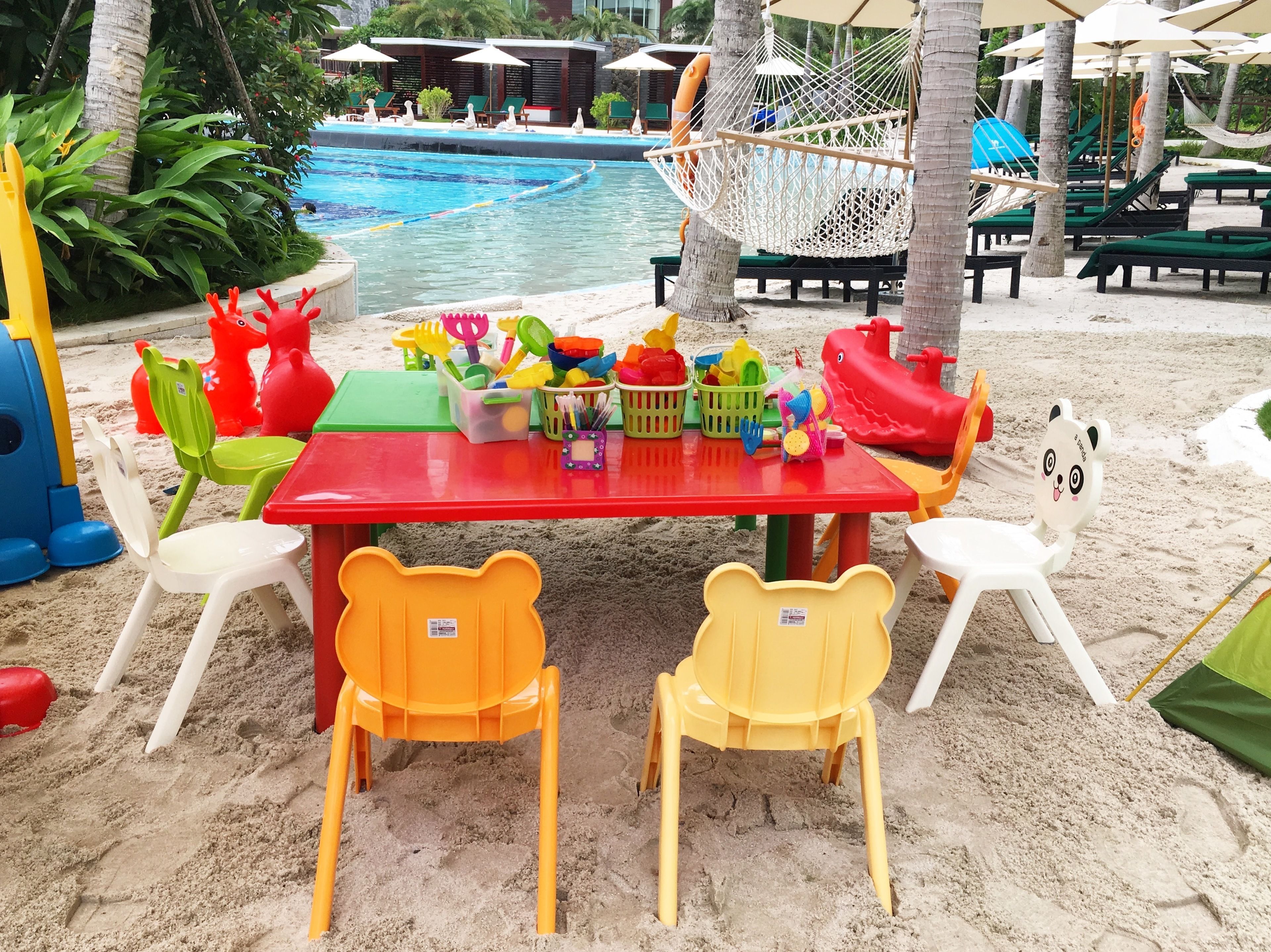 A beachside pool area with a red table, yellow chairs, and various beach toys.