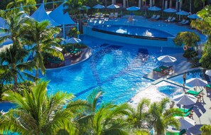 A large outdoor swimming pool with multiple lanes, surrounded by lounge chairs and umbrellas, and palm trees.