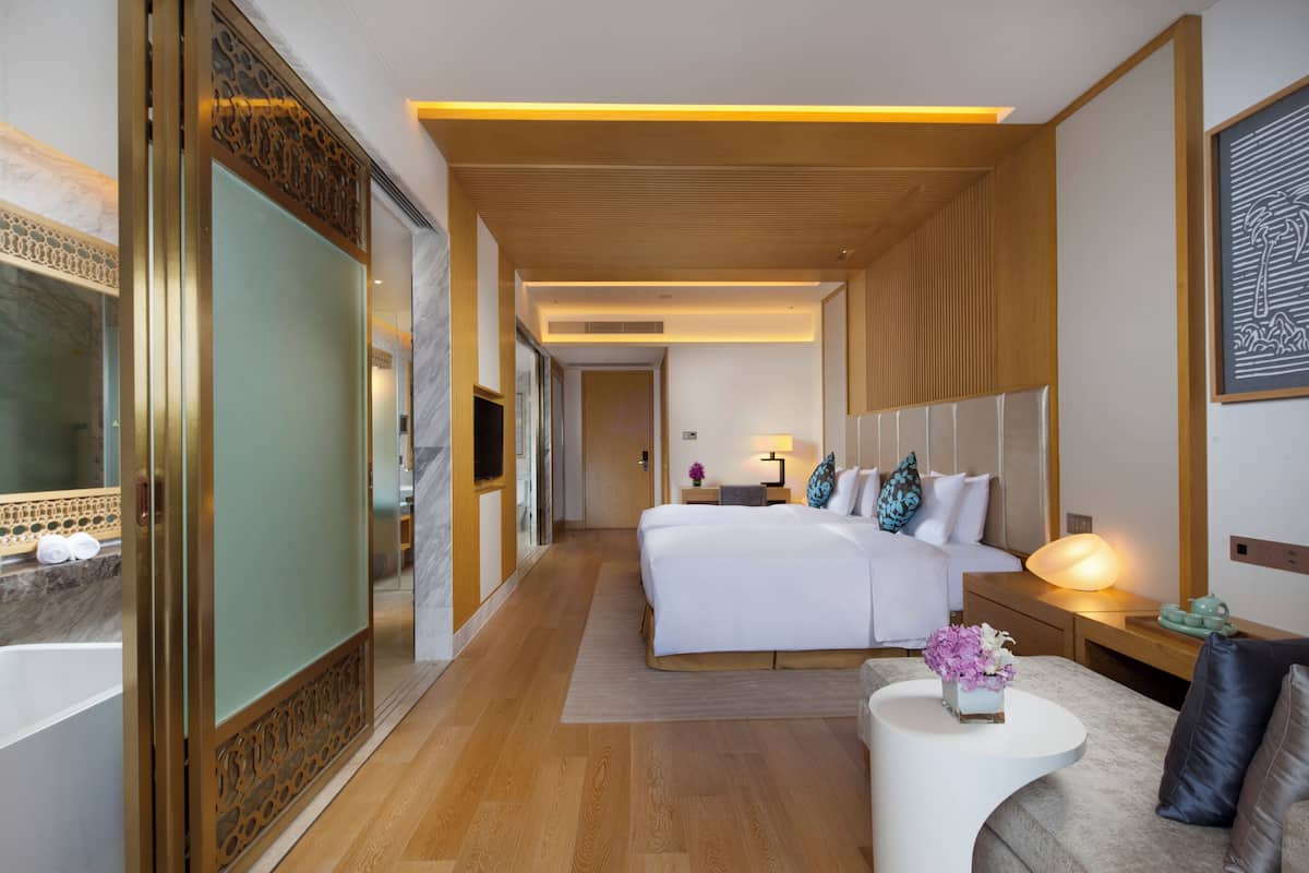 A modern hotel room with a large bed, a sitting area with a coffee table, and a bathroom with a large mirror and a bathtub.