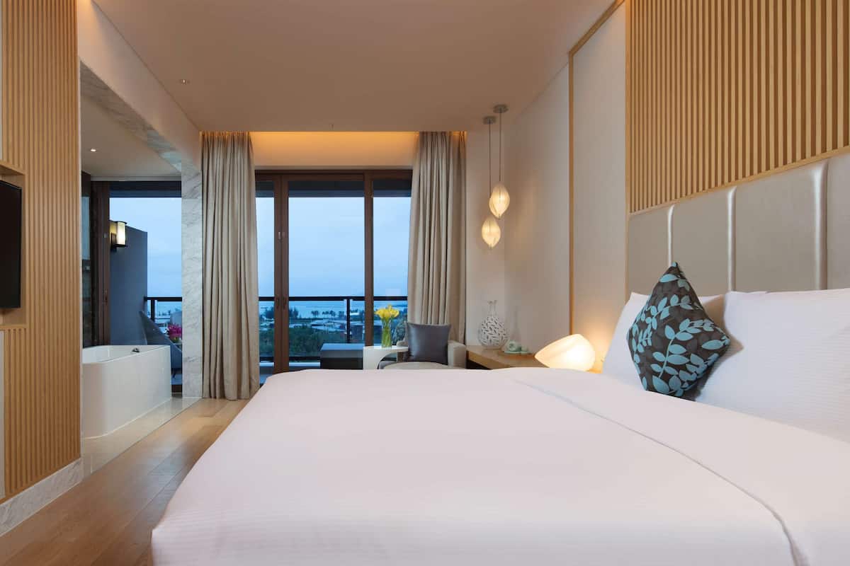 A hotel room with a large bed, a bedside table with a lamp, a view of the city through large windows, and a balcony with a view of the sea.
