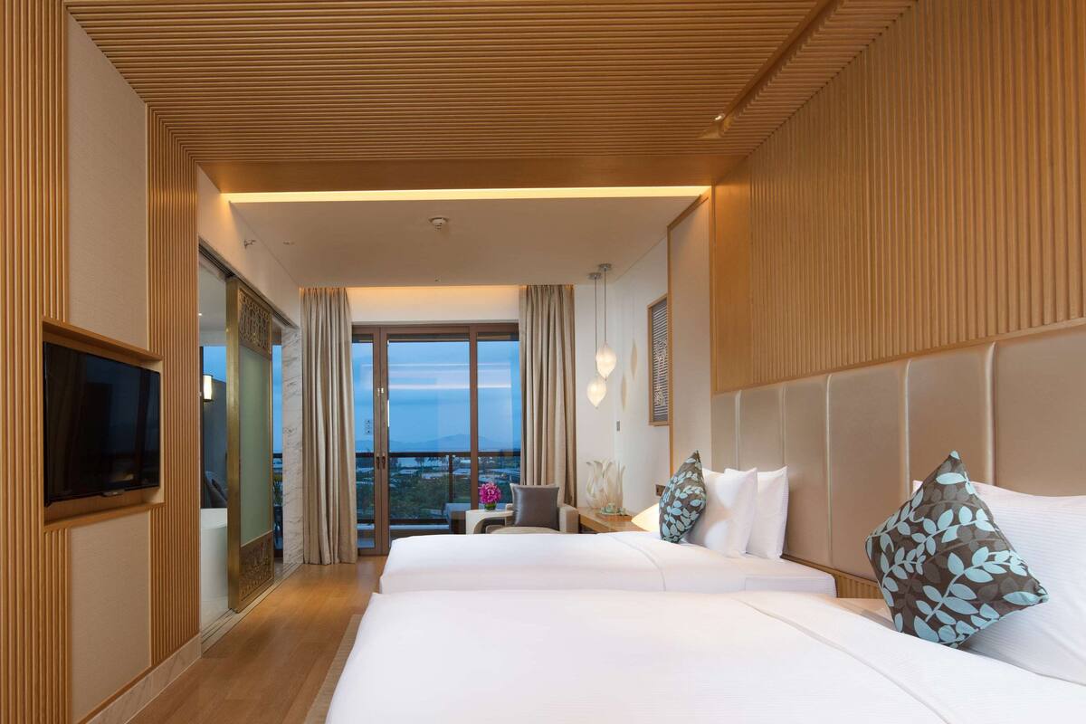A hotel room with two beds, a TV, a balcony with a view, and wooden paneling on the walls.
