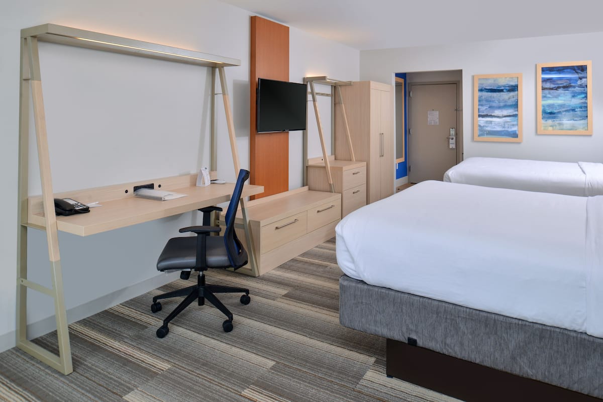 A hotel room with a desk, chair, bed, wardrobe, and artwork on the wall.